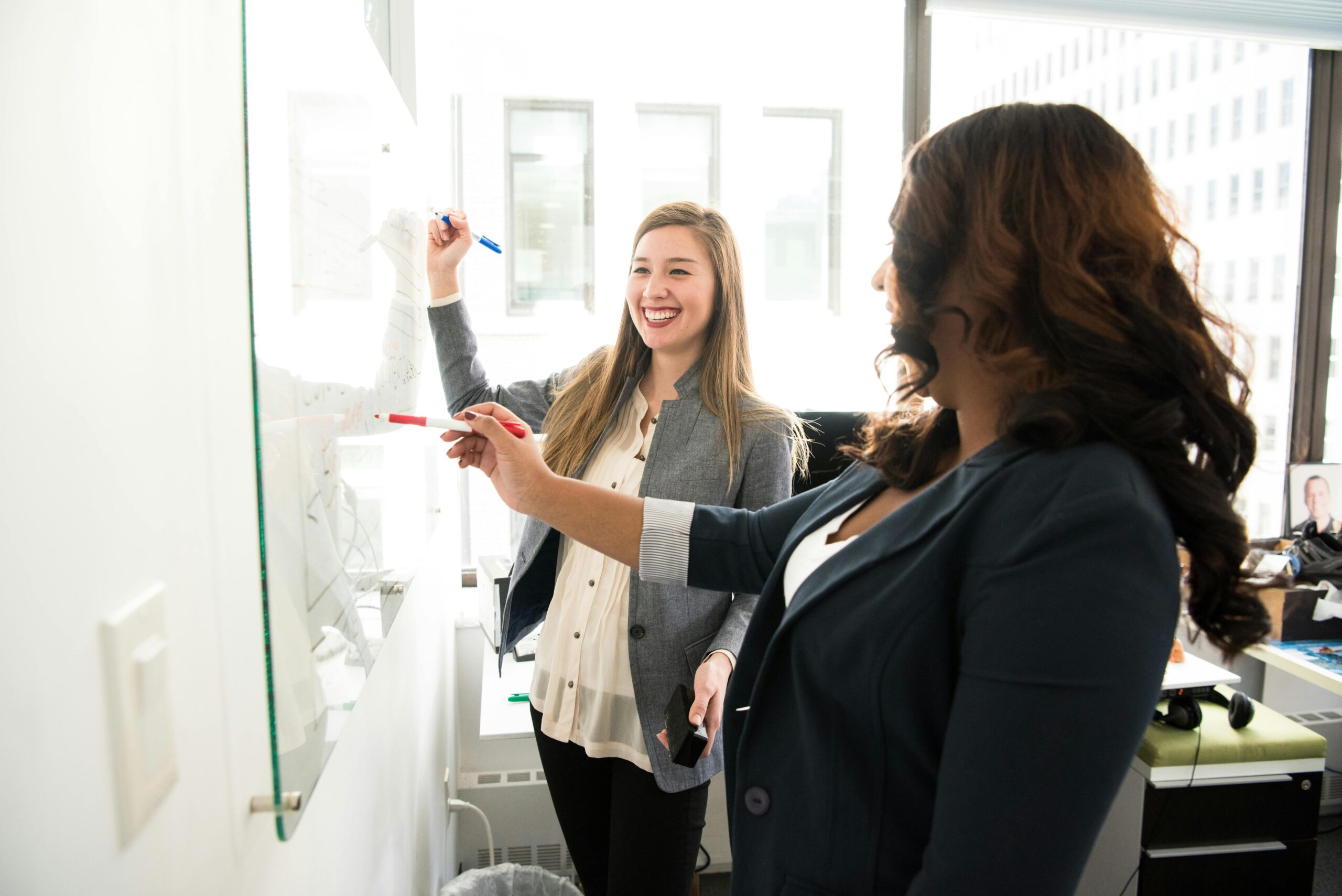 Two women writing on a dry-erase board.